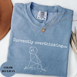 K&ouml;nnte beinhalten: Blaues Jeans-T-Shirt mit dem Text "Currently overthinking..." und einer Strichm&auml;nnchen-Grafik. Eine goldene Halskette mit Anh&auml;nger liegt auf dem Shirt. Das Shirt ist gefaltet und hat ein Comfort Colors-Etikett.