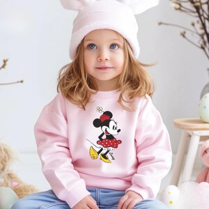Puede incluir: Una ni&ntilde;a lleva una sudadera rosa claro con un estampado de Minnie Mouse. La sudadera tiene mangas largas y cuello redondo. La ni&ntilde;a tambi&eacute;n lleva un gorro blanco con orejas de conejo y vaqueros azules. El estampado de Minnie Mouse tiene una falda roja con lunares blancos.