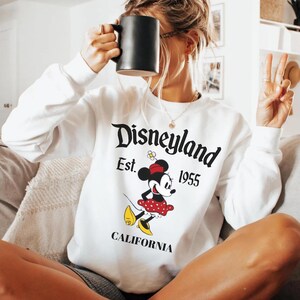 May include: White long-sleeve sweatshirt with a Disneyland graphic featuring Minnie Mouse. The design includes the text "Disneyland Est. 1955 CALIFORNIA" in black. Minnie Mouse is depicted in a red polka dot skirt and yellow shoes.