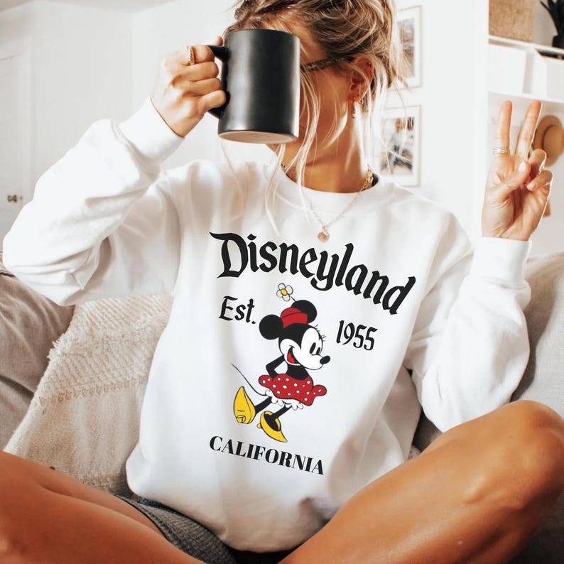 May include: White long-sleeve sweatshirt with a Disneyland graphic featuring Minnie Mouse. The design includes the text "Disneyland Est. 1955 CALIFORNIA" in black. Minnie Mouse is depicted in a red polka dot skirt and yellow shoes.