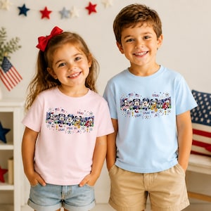 Pu&ograve; includere: Due bambini indossano magliette dai colori chiari con una grafica a cartoni animati. Le magliette presentano un disegno di personaggi dei cartoni animati, palloni da calcio e bandiere americane, con la parola "USA" ripetuta. La maglietta della ragazza &egrave; rosa e quella del ragazzo azzurra.