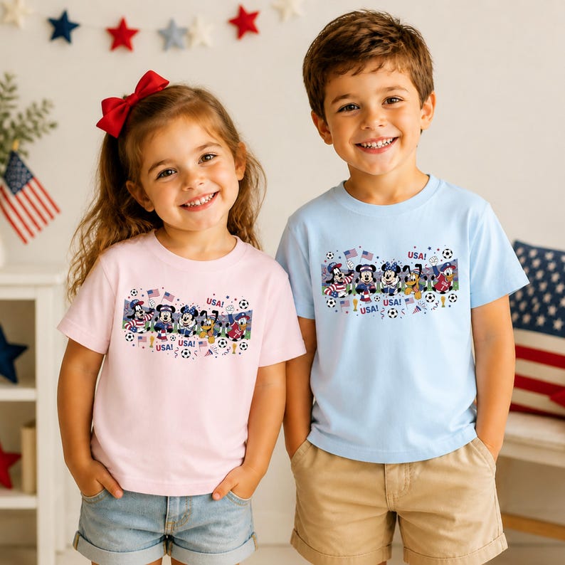 Pu&ograve; includere: Due bambini indossano magliette dai colori chiari con una grafica a cartoni animati. Le magliette presentano un disegno di personaggi dei cartoni animati, palloni da calcio e bandiere americane, con la parola "USA" ripetuta. La maglietta della ragazza &egrave; rosa e quella del ragazzo azzurra.
