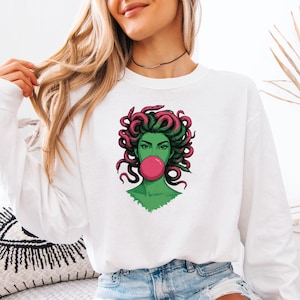 May include: White long-sleeve sweatshirt featuring a Medusa graphic. The illustration depicts Medusa with green skin, pink hair, and a bubblegum bubble. The design is a bold, colorful statement piece.