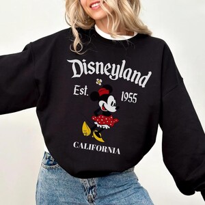May include: Black sweatshirt featuring the Disneyland logo, Minnie Mouse graphic, and the text "CALIFORNIA". The sweatshirt has a white collar and long sleeves. The Disneyland logo is in white, with the year "1955" below the logo.