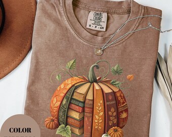 Könnte beinhalten: Espressofarbenes T-Shirt mit einem Kürbisdesign aus Büchern. Die Kürbisgrafik zeigt einen Stapel Bücher in der Mitte mit orangefarbenen und braunen Einbänden. Das Shirt hat einen Rundhalsausschnitt und kurze Ärmel. Eine silberne Halskette mit Anhänger ist sichtbar.