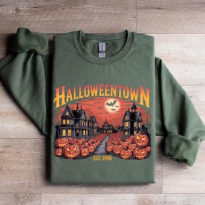May include: Green sweatshirt with a Halloween graphic. The design features a spooky town scene with jack-o'-lanterns, haunted houses, and the text "Halloweentown". The graphic also includes a full moon and bats. The text "EST. 1998" is also included.