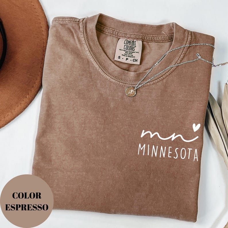 K&ouml;nnte beinhalten: Espressofarbenes T-Shirt mit dem Wort "Minnesota" in wei&szlig;er Schrift. Eine silberne Halskette mit einem kleinen Anh&auml;nger liegt auf dem Shirt. Das Shirt hat einen Rundhalsausschnitt und kurze &Auml;rmel. Das Shirt ist in Gr&ouml;&szlig;e S.