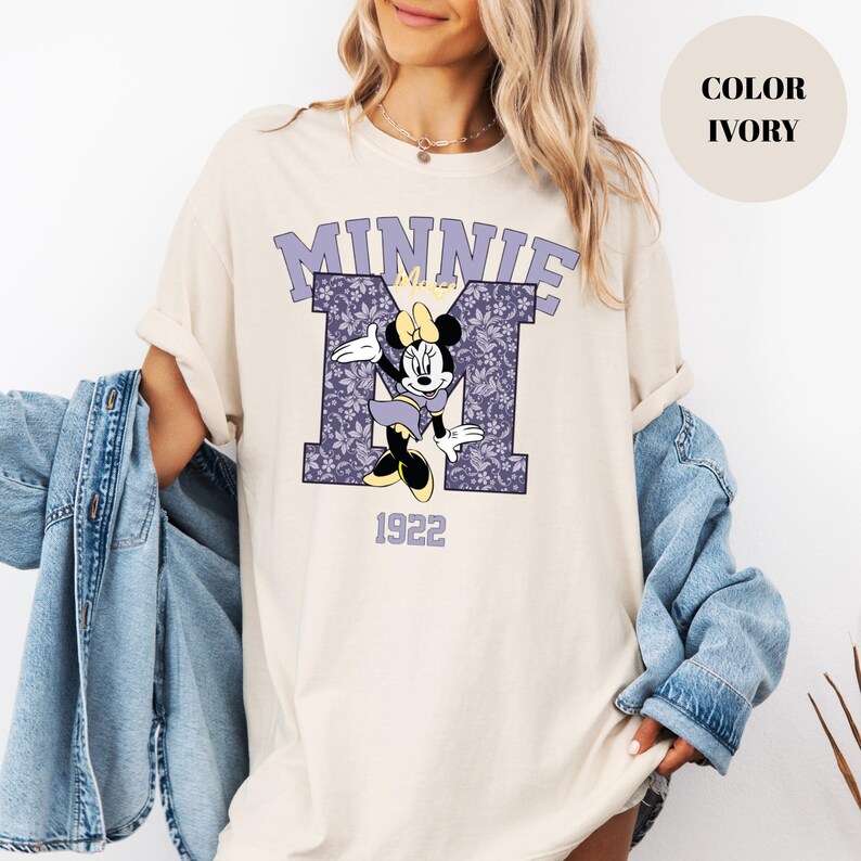 Pu&ograve; includere: T-shirt color avorio con grafica di Minnie Mouse. Il design include la parola "MINNIE" in lavanda, una "M" a motivi floreali e l'anno "1922". Minnie Mouse &egrave; raffigurata con un vestito e un fiocco viola.