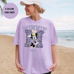 Pu&ograve; includere: T-shirt color orchidea con grafica di Minnie Mouse. La grafica include la scritta "MINNIE" sopra una "M" a motivi floreali con Minnie Mouse al centro e l'anno "1922" in basso. La maglietta &egrave; di colore viola chiaro.