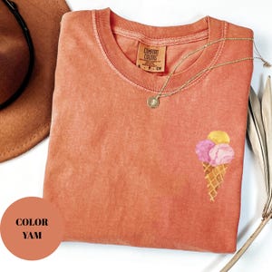 May include: A coral-colored t-shirt with a crew neck and short sleeves. The shirt features a watercolor illustration of an ice cream cone with three scoops. A gold necklace with a pendant is draped on the shirt. The tag reads "Comfort Colors".