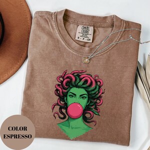 May include: Espresso-colored t-shirt featuring a graphic of a woman with green skin, pink hair, and a pink bubblegum bubble. The shirt has a crew neck and short sleeves. A silver necklace with a small pendant is draped across the shirt.
