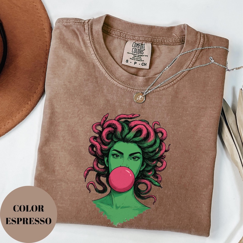 May include: Espresso-colored t-shirt featuring a graphic of a woman with green skin, pink hair, and a pink bubblegum bubble. The shirt has a crew neck and short sleeves. A silver necklace with a small pendant is draped across the shirt.