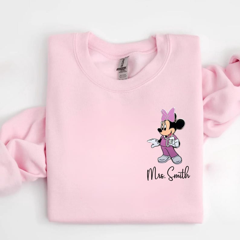 以下が含まれることがあります： 紫と白のミニーマウスのイラストが描かれた、明るいピンク色のスウェットシャツ。カップを持っています。「Mrs. Smith」の文字が漫画のキャラクターの下に黒い筆記体で印刷されています。