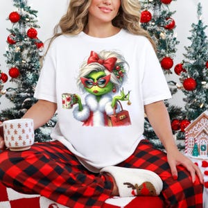 May include: White t-shirt featuring a cartoon character with green skin, a red bow, and sunglasses. The character is holding a mug and a red purse. The shirt is paired with red and black plaid pajama pants and a pair of reindeer slippers.