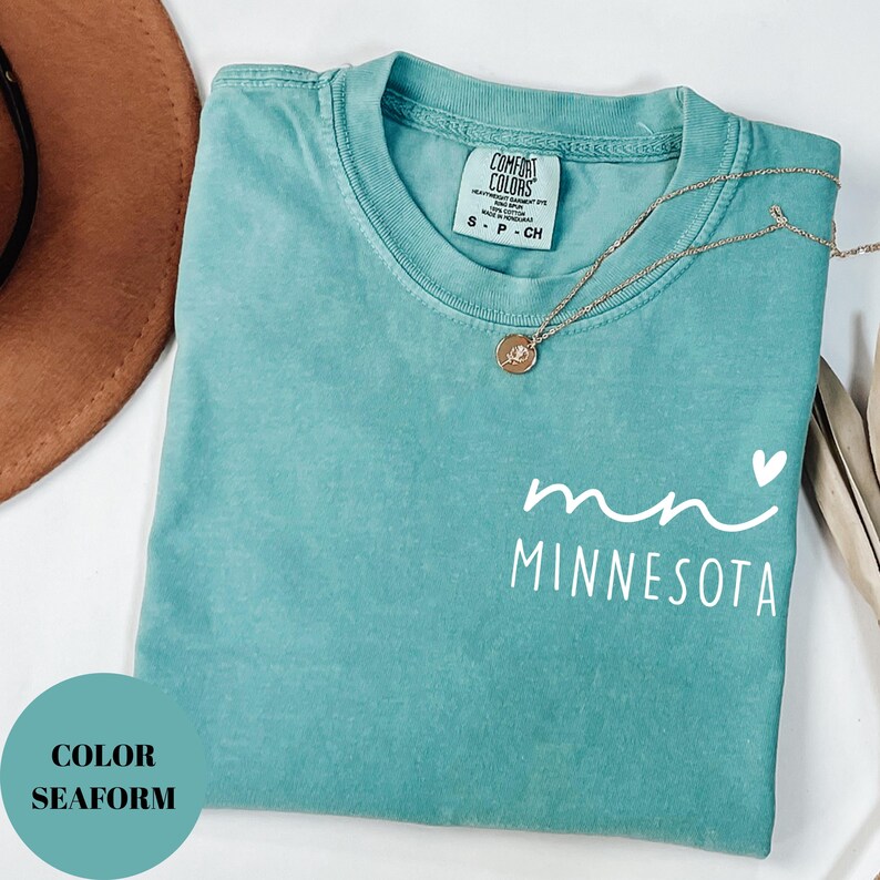 K&ouml;nnte beinhalten: T&uuml;rkisfarbenes T-Shirt mit dem wei&szlig;en Text "mn Minnesota". Eine goldene Halskette mit Anh&auml;nger liegt &uuml;ber dem Shirt. Das Shirt hat einen Rundhalsausschnitt und ein Etikett mit der Aufschrift "Comfort Colors".