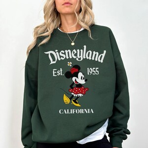 May include: Dark green Disneyland sweatshirt with Minnie Mouse in a red polka dot skirt and yellow shoes. The sweatshirt has the text "Disneyland Est. 1955 California" in white lettering. The sweatshirt is a crew neck style.