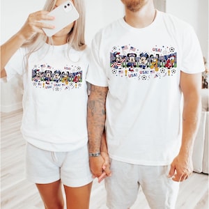 Pu&ograve; includere: T-shirt bianche con una grafica colorata raffigurante personaggi Disney in abiti patriottici, palloni da calcio e bandiere americane. La parola "USA!" &egrave; stampata sulle magliette. Le magliette sono indossate da due persone.