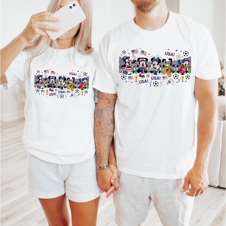 Pu&ograve; includere: T-shirt bianche con una grafica colorata raffigurante personaggi Disney in abiti patriottici, palloni da calcio e bandiere americane. La parola "USA!" &egrave; stampata sulle magliette. Le magliette sono indossate da due persone.