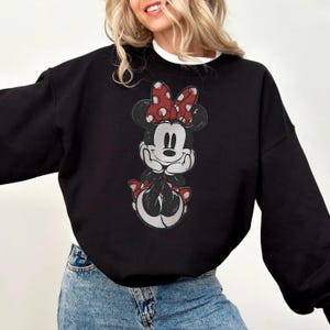 May include: Black sweatshirt featuring a graphic of Minnie Mouse. The cartoon character is depicted with a red and white polka dot bow, and red shoes. The sweatshirt has long sleeves and a crew neck.