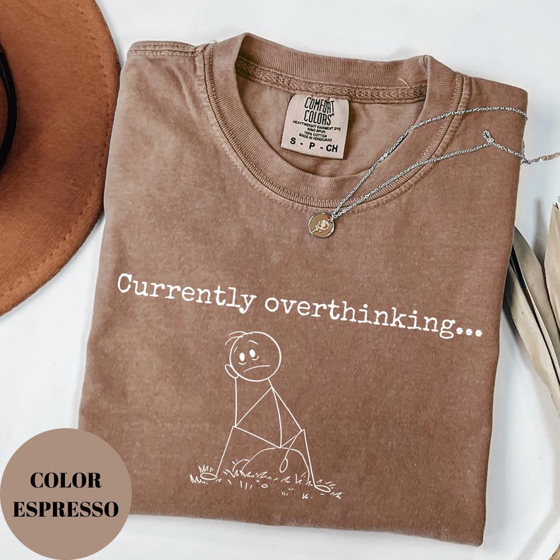 K&ouml;nnte beinhalten: Espressofarbenes T-Shirt mit dem Text "Currently overthinking..." und einer Strichm&auml;nnchen-Grafik. Das Shirt hat einen Rundhalsausschnitt und ist gefaltet. Eine silberne Halskette mit Anh&auml;nger liegt oben auf dem Shirt.