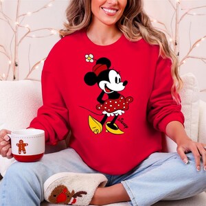 Puede incluir: Una sudadera roja con un gr&aacute;fico de Minnie Mouse. El personaje de dibujos animados lleva un vestido de lunares rojos, zapatos amarillos y un lazo rojo. La palabra "Disney" est&aacute; impresa en la parte inferior derecha del gr&aacute;fico.