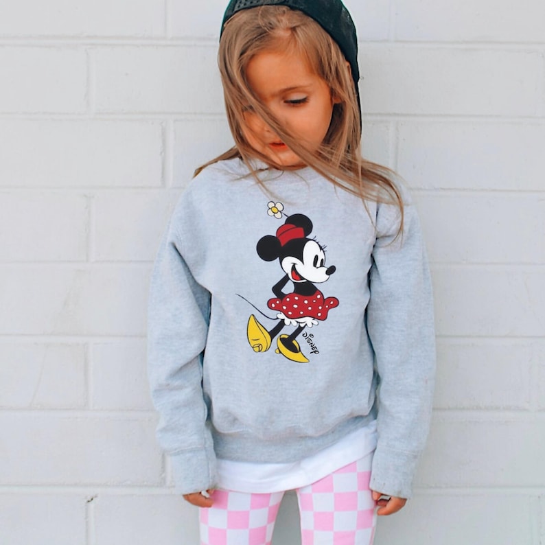 Puede incluir: Una ni&ntilde;a peque&ntilde;a con una sudadera gris con un dise&ntilde;o de Minnie Mouse. El dise&ntilde;o muestra a Minnie con un vestido rojo de lunares y zapatos amarillos. La ni&ntilde;a tambi&eacute;n lleva leggings a cuadros rosas y blancos y una gorra de b&eacute;isbol negra.
