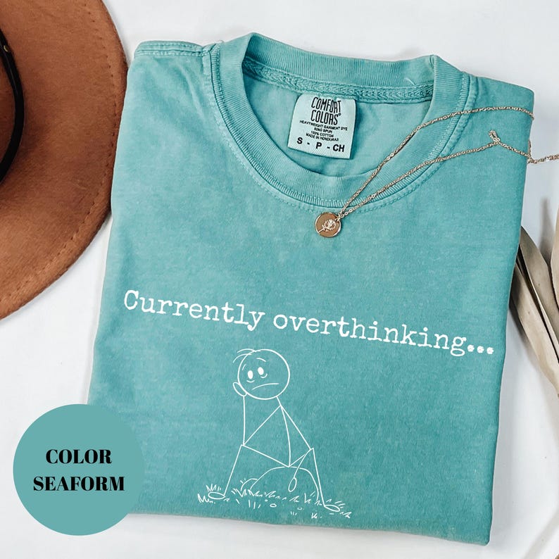 K&ouml;nnte beinhalten: T&uuml;rkisgr&uuml;nes T-Shirt mit dem Text "Currently overthinking..." und einer Strichm&auml;nnchen-Grafik. Das Shirt hat einen Rundhalsausschnitt und kurze &Auml;rmel. Eine goldene Halskette mit einem kleinen Anh&auml;nger liegt &uuml;ber dem Shirt.