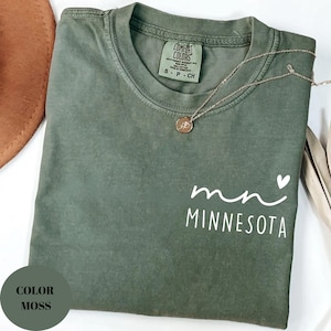 K&ouml;nnte beinhalten: Ein moosgr&uuml;nes T-Shirt mit dem Wort "Minnesota" in Wei&szlig; aufgedruckt. Eine goldene Halskette mit Anh&auml;nger liegt auf dem Shirt. Das Shirt hat einen Rundhalsausschnitt und kurze &Auml;rmel. Das Shirt ist gefaltet.