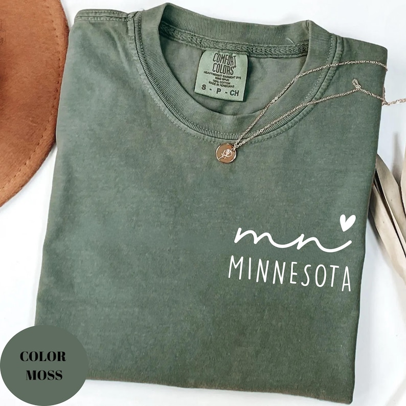 K&ouml;nnte beinhalten: Ein moosgr&uuml;nes T-Shirt mit dem Wort "Minnesota" in Wei&szlig; aufgedruckt. Eine goldene Halskette mit Anh&auml;nger liegt auf dem Shirt. Das Shirt hat einen Rundhalsausschnitt und kurze &Auml;rmel. Das Shirt ist gefaltet.