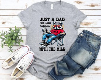 Pode incluir: Uma camiseta cinza mesclada com o texto "JUST A DAD WHO ALWAYS CAME BACK WITH THE MILK". O gráfico apresenta um guaxinim usando óculos de sol e uma camisa vermelha, sentado em uma cadeira e segurando uma caixa de leite. Tênis Converse brancos e jeans azuis também estão na imagem.