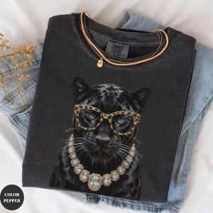 May include: A charcoal gray t-shirt featuring a graphic of a black panther wearing leopard print glasses and a large, multi-faceted crystal statement necklace. The shirt has a crew neckline and a Comfort Colors brand label on the inner collar.