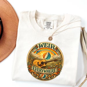 May include: Off-white t-shirt with a circular graphic that reads "WEIR EVERYWHERE." The design includes a guitar, rainbow, mountains, and mushrooms. The shirt has a Comfort Colors label.