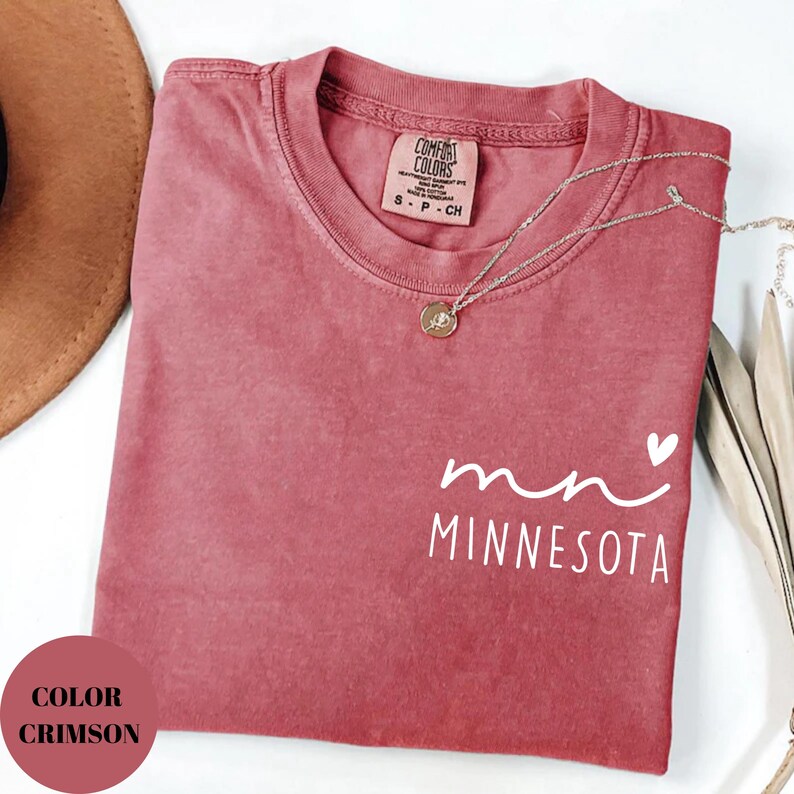 K&ouml;nnte beinhalten: Karmesinrotes T-Shirt mit dem Wort "Minnesota" in wei&szlig;er Schrift, einem kleinen Herz und der Abk&uuml;rzung "mn". Das Shirt hat einen Rundhalsausschnitt und ein Comfort Colors-Etikett. Eine silberne Halskette mit Anh&auml;nger liegt auf dem Shirt.