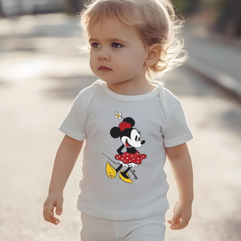 Puede incluir: Body de beb&eacute; blanco con un gr&aacute;fico de Minnie Mouse. El personaje de dibujos animados est&aacute; representado con un vestido de lunares rojos y blancos, zapatos amarillos y un lazo rojo. El logotipo de Disney est&aacute; impreso debajo.