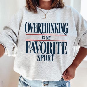 Pode incluir: Uma camisola de gola redonda cinzento-claro com o texto azul-escuro OVERTHINKING IS MY FAVORITE SPORT, com duas linhas horizontais vermelhas finas a separar as palavras. A peça tem mangas compridas, gola canelada e é usada com calças de ganga.