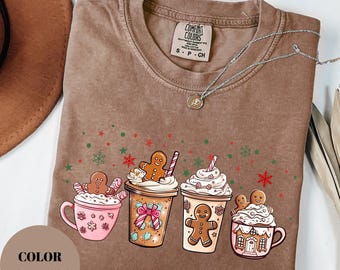 Puede incluir: Camiseta color espresso con un diseño festivo de hombre de jengibre y tazas de café. La camiseta tiene cuello redondo y mangas cortas. El gráfico incluye hombres de jengibre, crema batida y detalles navideños. El texto "COLOR ESPRESSO" es visible.