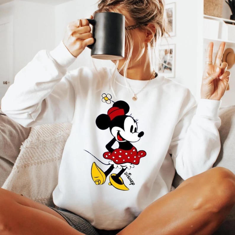 Puede incluir: Sudadera blanca con un gr&aacute;fico de Minnie Mouse. El personaje de dibujos animados lleva una falda roja de lunares, zapatos amarillos y un lazo rojo. El logotipo de Disney est&aacute; impreso debajo. La sudadera tiene mangas largas.