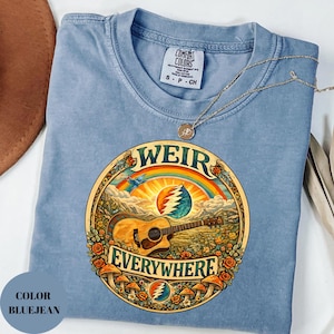 May include: Bluejean Comfort Colors t-shirt with a circular graphic featuring a guitar, rainbow, and the words "WEIR EVERYWHERE." The design includes mushrooms and floral elements. A gold necklace rests on the shirt.