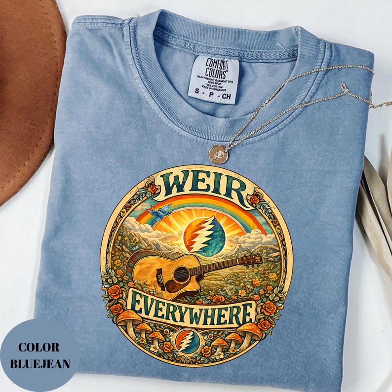 May include: Bluejean Comfort Colors t-shirt with a circular graphic featuring a guitar, rainbow, and the words "WEIR EVERYWHERE." The design includes mushrooms and floral elements. A gold necklace rests on the shirt.
