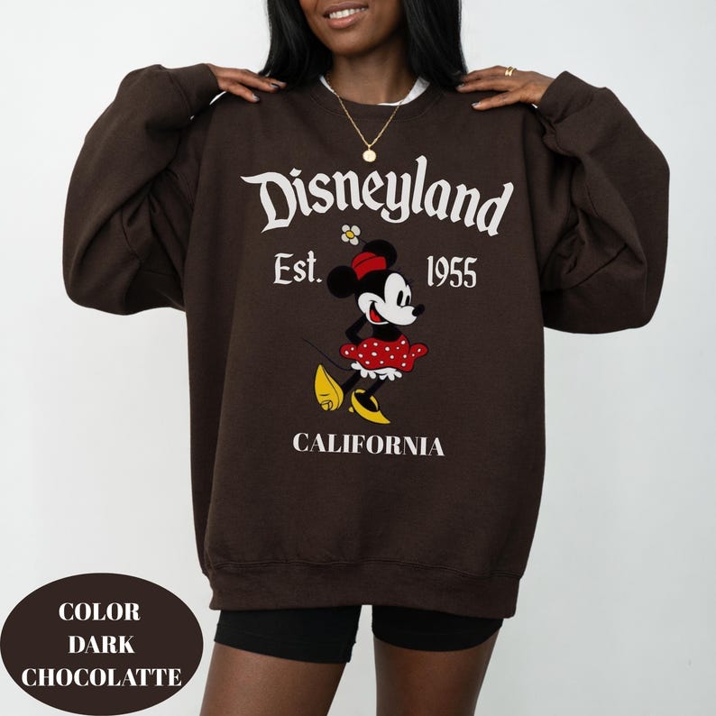 May include: A dark chocolate-coloured sweatshirt with the Disneyland logo, the text "Est. 1955", and a Minnie Mouse graphic. The word "CALIFORNIA" is printed below the graphic. The sweatshirt has a relaxed fit.