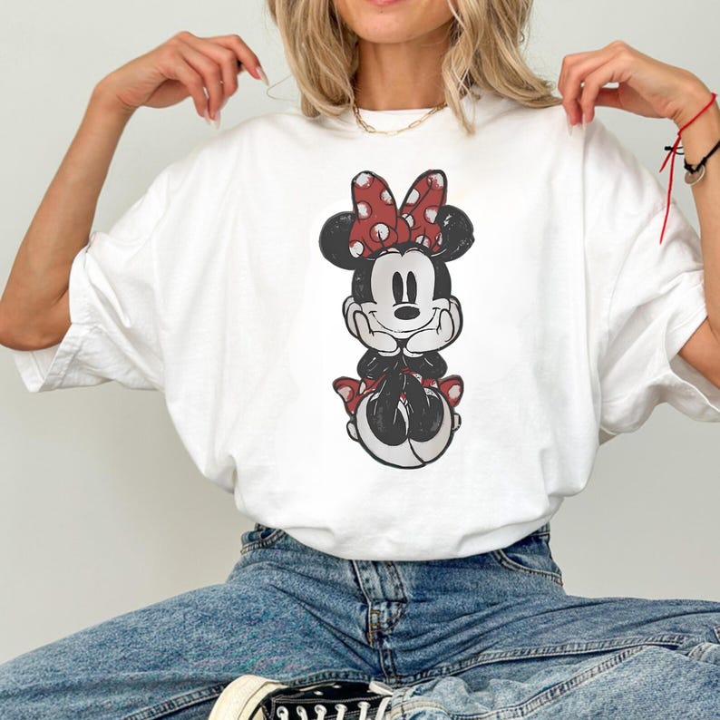 May include: White t-shirt featuring a distressed graphic of Minnie Mouse. Minnie is depicted with a red polka dot bow, black ears, and a red polka dot skirt. The shirt is made of a soft, comfortable material.