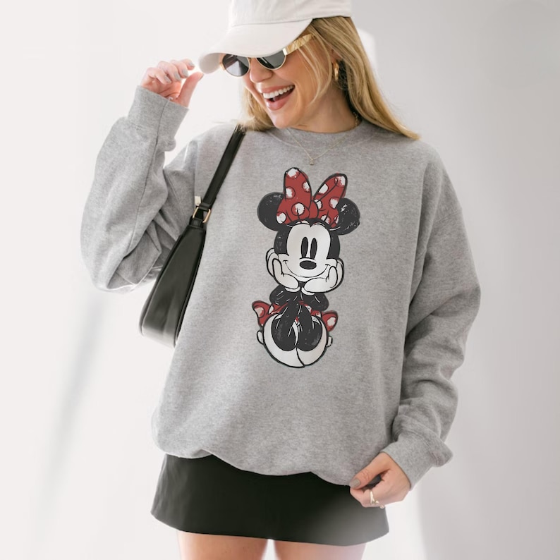 May include: A gray sweatshirt featuring a distressed graphic of Minnie Mouse with a red bow and accents. The sweatshirt is long-sleeved and has a crew neck. The overall style is casual and comfortable.