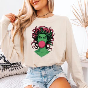 May include: Cream-colored long-sleeve shirt featuring a graphic of Medusa with green skin, pink hair, and a pink bubble gum bubble. The graphic is centered on the shirt. The model is wearing denim shorts.