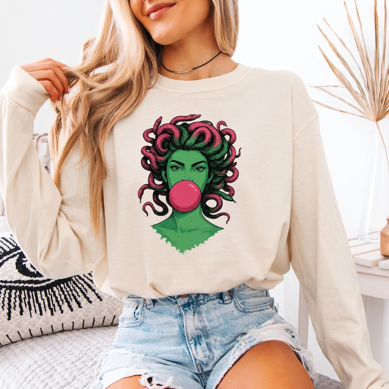 May include: Cream-colored long-sleeve shirt featuring a graphic of Medusa with green skin, pink hair, and a pink bubble gum bubble. The graphic is centered on the shirt. The model is wearing denim shorts.