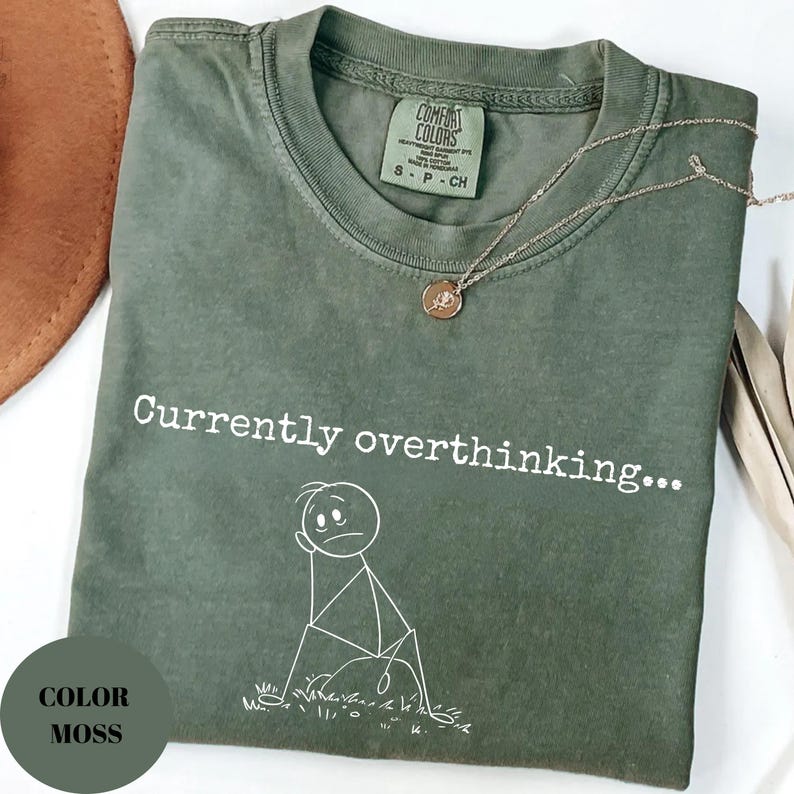 K&ouml;nnte beinhalten: Ein moosgr&uuml;nes T-Shirt mit dem Schriftzug "Currently overthinking..." und einer Strichm&auml;nnchen-Grafik. Eine goldene Halskette mit Anh&auml;nger liegt auf dem Shirt. Das Shirt hat ein Comfort Colors-Etikett.