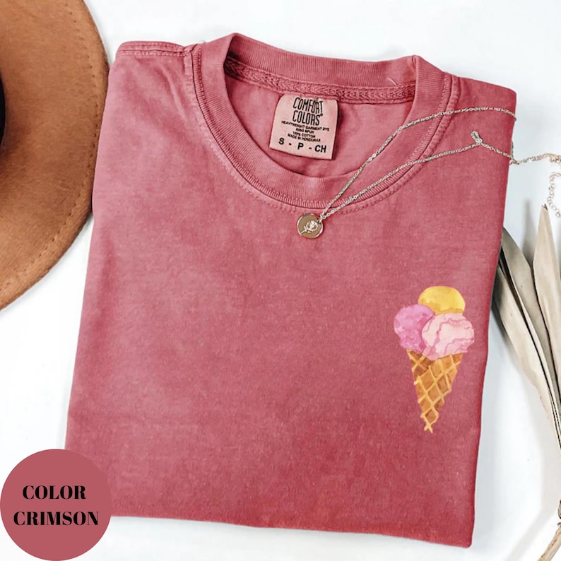 May include: Crimson-colored t-shirt with a crew neck and short sleeves. The shirt features an embroidered ice cream cone design with three scoops in pink and yellow. A silver necklace with a pendant rests on the shirt.