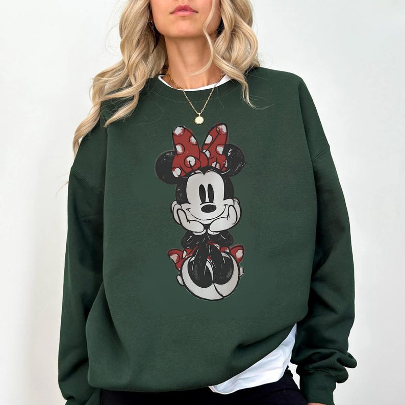 May include: Dark green sweatshirt featuring a distressed graphic of Minnie Mouse. Minnie is depicted in black and white with a red polka dot bow and matching accents. The sweatshirt has a white crew neck collar.