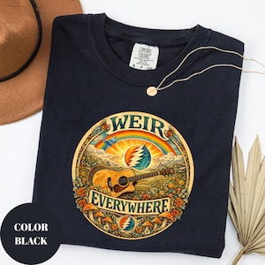 May include: Black t-shirt with a circular graphic featuring a guitar, rainbow, and the words "WEIR EVERYWHERE." The design includes a mountain landscape, flowers, and mushrooms. A gold necklace and a brown hat are also visible.