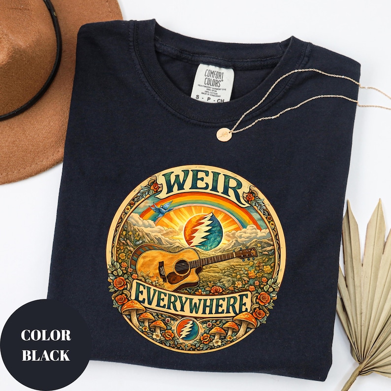 May include: Black t-shirt with a circular graphic featuring a guitar, rainbow, and the words "WEIR EVERYWHERE." The design includes a mountain landscape, flowers, and mushrooms. A gold necklace and a brown hat are also visible.