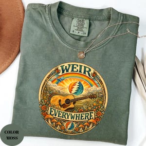 May include: A moss green t-shirt with a circular graphic featuring a guitar, rainbow, and the words "WEIR EVERYWHERE." The design includes mushrooms, roses, and a stylized sun. A silver necklace with a pendant rests on the shirt.